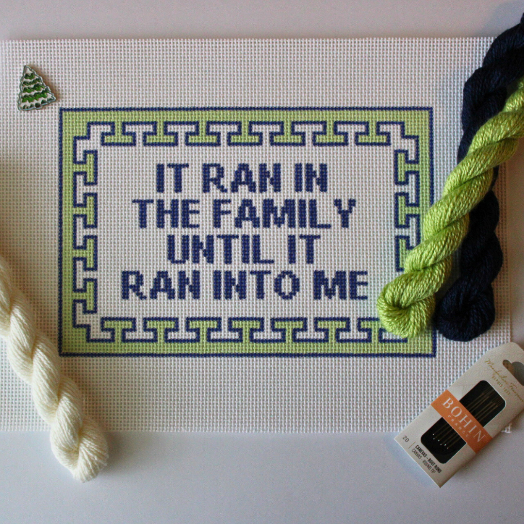 Threaded & Tipsy hand-painted needlepoint canvas featuring the phrase “IT RAN IN THE FAMILY UNTIL IT RAN INTO ME” in bold navy blue block letters. The quote is framed by a navy and lime green geometric border. Surrounding the canvas are twisted skeins of needlepoint threads in cream, lime green, and navy blue, along with a pack of Bohin embroidery needles in the bottom right corner. A small enamel pin shaped like a green Christmas tree is pinned in the upper left corner of the canvas.