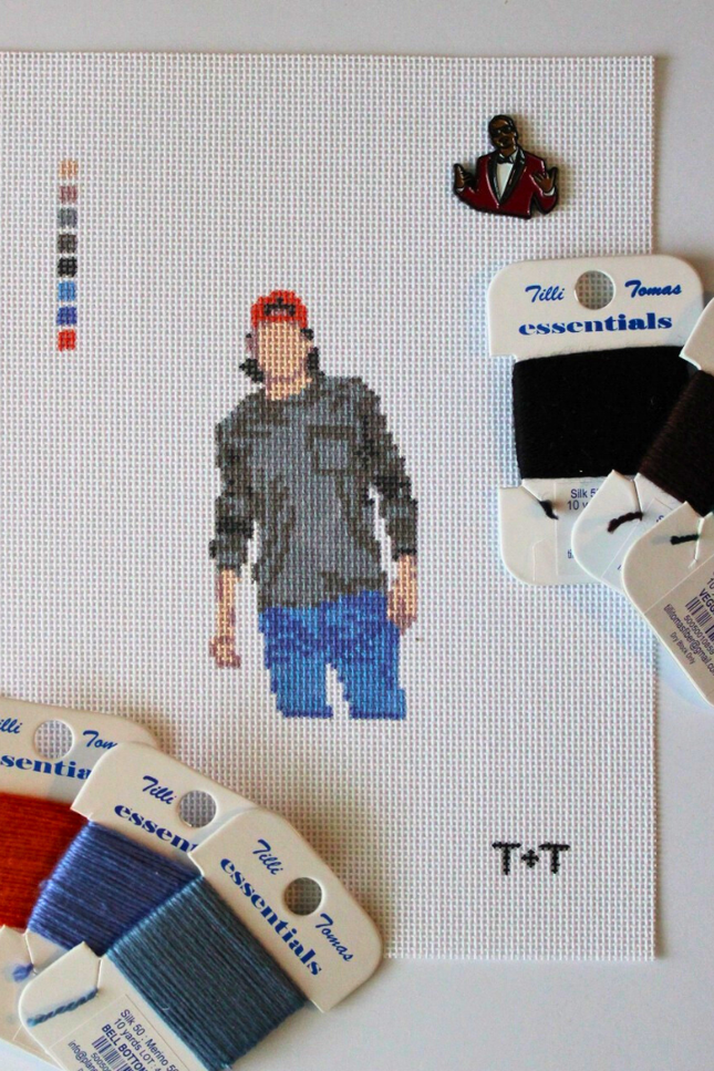 Alt text:
Threaded & Tipsy needlepoint canvas featuring a pixel-style figure of a man wearing a red baseball cap, gray shirt, and blue jeans. The canvas includes a stitched color guide and the initials "T+T" in the bottom right corner. Surrounding the canvas are six spools of Threaded & Tipsy needlepoint threads in black, brown, green, orange, and two shades of blue. A small enamel pin of a man in a red blazer is pinned near the top of the canvas.