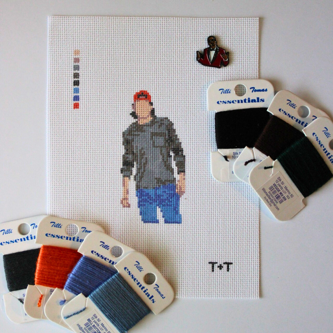 Alt text:
Threaded &amp; Tipsy needlepoint canvas featuring a pixel-style figure of a man wearing a red baseball cap, gray shirt, and blue jeans. The canvas includes a stitched color guide and the initials "T+T" in the bottom right corner. Surrounding the canvas are six spools of Threaded &amp; Tipsy needlepoint threads in black, brown, green, orange, and two shades of blue. A small enamel pin of a man in a red blazer is pinned near the top of the canvas.