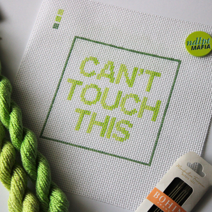 Threaded &amp; Tipsy hand-painted needlepoint canvas featuring the phrase “CAN’T TOUCH THIS” in bold gradient lime and yellow-green block letters, framed by a green rectangular border. A small painted color guide with three green shades appears in the top left corner. Surrounding the canvas are twisted skeins of green needlepoint threads, a pack of Bohin embroidery needles in the bottom right, and a round lime green enamel pin in the top right corner that reads “ndlpt MAFIA.” 