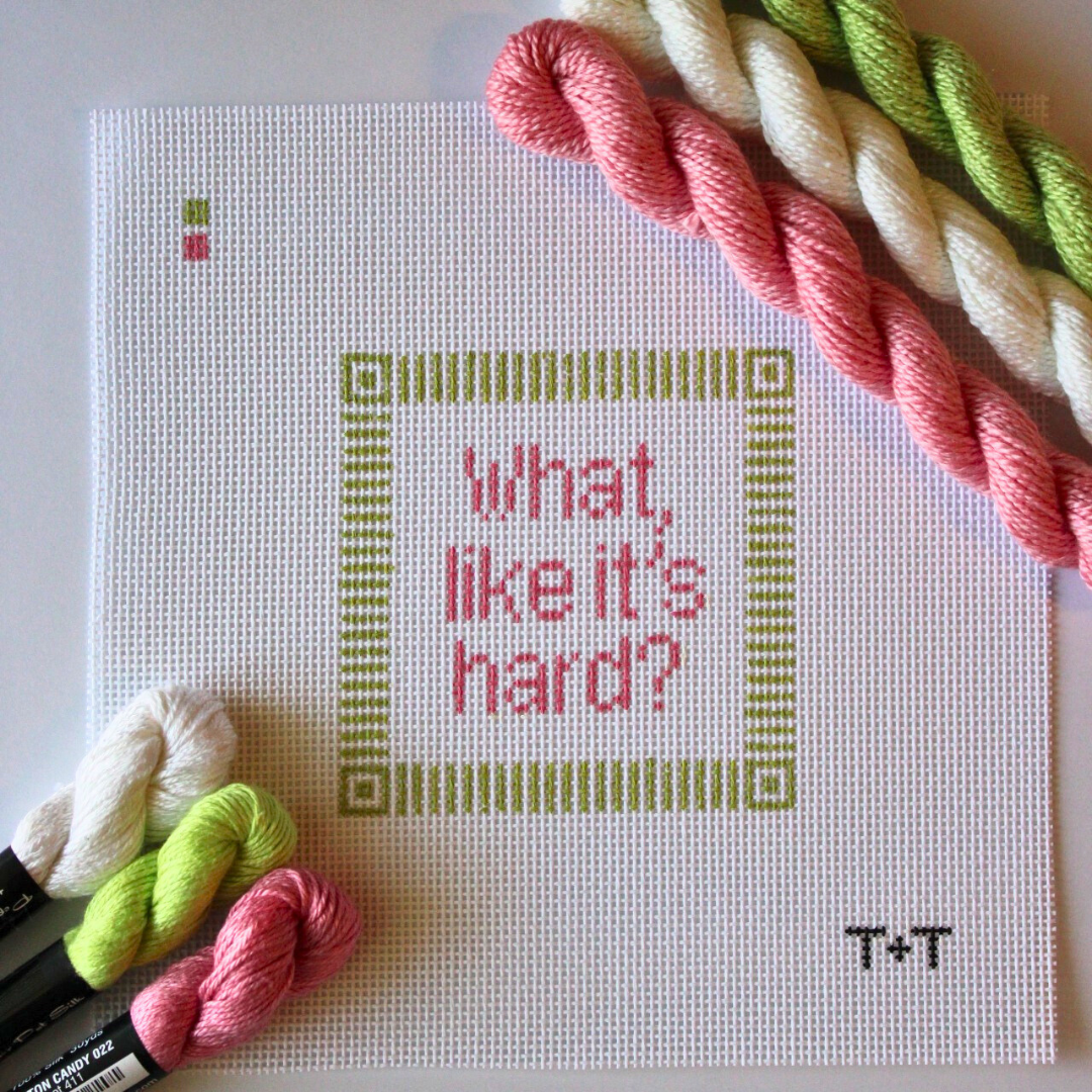 Threaded &amp; Tipsy hand-painted needlepoint canvas featuring the phrase “what, like it’s hard?” in pink lettering, centered within a green geometric border. The initials "T+T" are painted in black in the bottom right corner. Three skeins of Threaded &amp; Tipsy needlepoint threads in white, lime green, and pink are placed beside and above the canvas. A small painted color guide appears in the upper left corner.