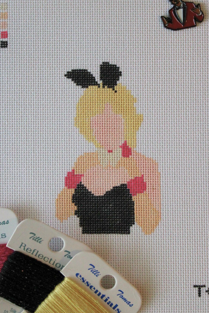 Threaded & Tipsy hand-painted needlepoint canvas featuring a faceless figure of a blonde woman in a black strapless outfit with pink gloves, a white collar, and black bunny ears. A painted color guide is visible in the upper left corner, and the initials "T+T" are painted in black in the bottom right corner. Surrounding the canvas are spools of Threaded & Tipsy needlepoint threads in pink, black, and yellow. A small enamel pin of a man in a red blazer is pinned near the top of the canvas.