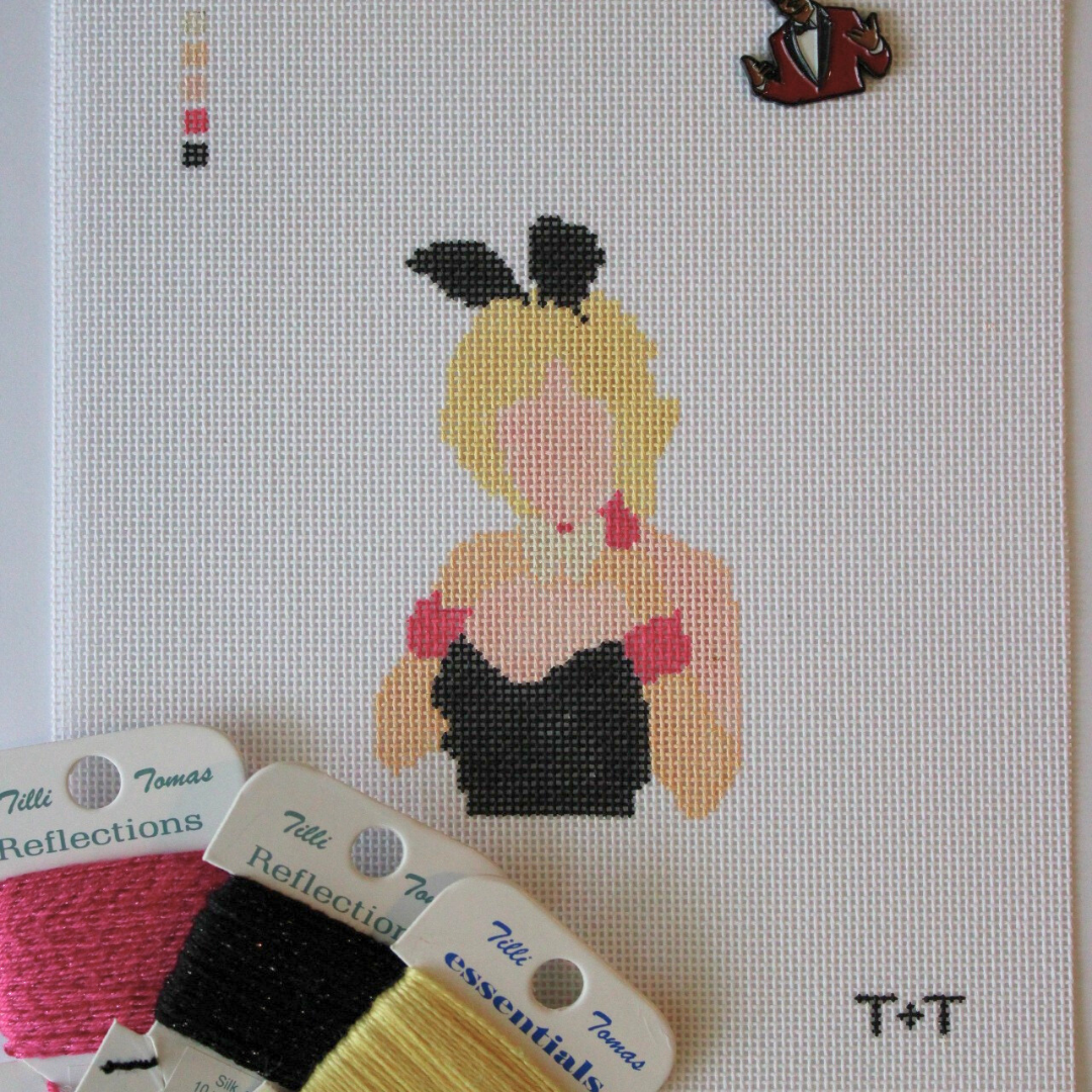 Threaded &amp; Tipsy hand-painted needlepoint canvas featuring a faceless figure of a blonde woman in a black strapless outfit with pink gloves, a white collar, and black bunny ears. A painted color guide is visible in the upper left corner, and the initials "T+T" are painted in black in the bottom right corner. Surrounding the canvas are spools of Threaded &amp; Tipsy needlepoint threads in pink, black, and yellow. A small enamel pin of a man in a red blazer is pinned near the top of the canvas.