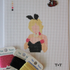 Threaded & Tipsy hand-painted needlepoint canvas featuring a faceless figure of a blonde woman in a black strapless outfit with pink gloves, a white collar, and black bunny ears. A painted color guide is visible in the upper left corner, and the initials "T+T" are painted in black in the bottom right corner. Surrounding the canvas are spools of Threaded & Tipsy needlepoint threads in pink, black, and yellow. A small enamel pin of a man in a red blazer is pinned near the top of the canvas.