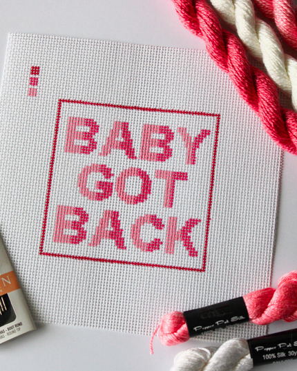 Alt text:
Threaded & Tipsy hand-painted needlepoint canvas featuring the phrase “BABY GOT BACK” in bold pink and red gradient block letters, centered within a red rectangular border. A small painted color guide with four shades of pink and red appears in the upper left corner. Surrounding the canvas are twisted skeins of pink, red, and white needlepoint threads, a pack of Bohin embroidery needles in the bottom left, and additional silk threads in the bottom right. The canvas is unstitched and laid flat on a