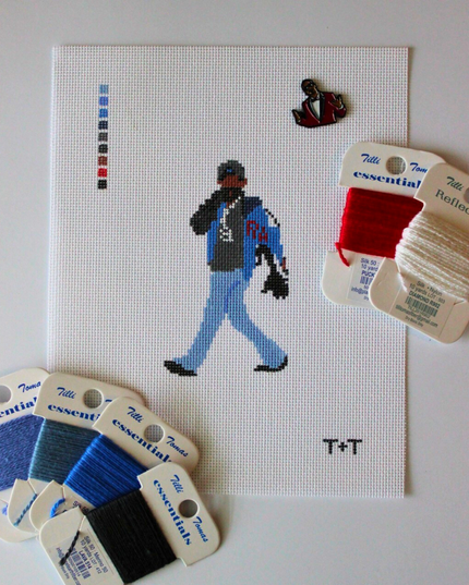 Threaded & Tipsy hand-painted needlepoint canvas featuring a pixel-style figure of a man walking while holding a phone to his ear, dressed in a black cap, black hoodie, blue jeans, and a blue and black jacket with red “RM” lettering. A painted color guide is visible on the upper left side, and the initials "T+T" are painted in black in the bottom right corner. Surrounding the canvas are Threaded & Tipsy needlepoint threads in shades of blue, black, red, and shimmery white. A small enamel pin of a man 