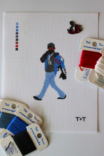 Threaded & Tipsy hand-painted needlepoint canvas featuring a pixel-style figure of a man walking while holding a phone to his ear, dressed in a black cap, black hoodie, blue jeans, and a blue and black jacket with red “RM” lettering. A painted color guide is visible on the upper left side, and the initials "T+T" are painted in black in the bottom right corner. Surrounding the canvas are Threaded & Tipsy needlepoint threads in shades of blue, black, red, and shimmery white. A small enamel pin of a man 