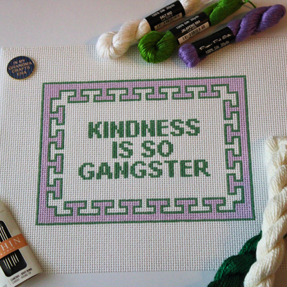 Threaded &amp; Tipsy hand-painted needlepoint canvas featuring the phrase “KINDNESS IS SO GANGSTER” in bold green block letters, framed by a lavender and green geometric border. The canvas is surrounded by silk needlepoint threads in white, green, and purple, along with a pack of thin needles in the bottom left and a circular enamel pin that reads “MY GRANDMA CRAFTS” in the top left corner. Partially visible twisted hanks of thread rest along the top and right side of the canvas.