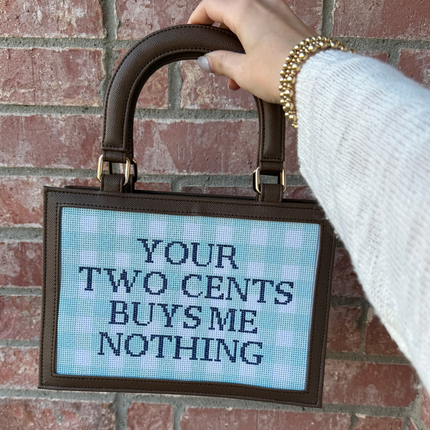 Hand holding a self finishing needlepoint bag with a digital display saying 'Your two cents buys me nothing' against a brick wall. 