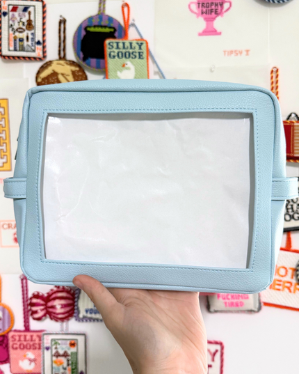 Light blue self finishing needlepoint pouch held by a hand against a colorful background with various small objects and text.