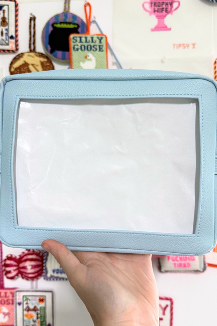 Light blue self finishing needlepoint pouch held by a hand against a colorful background with various small objects and text.