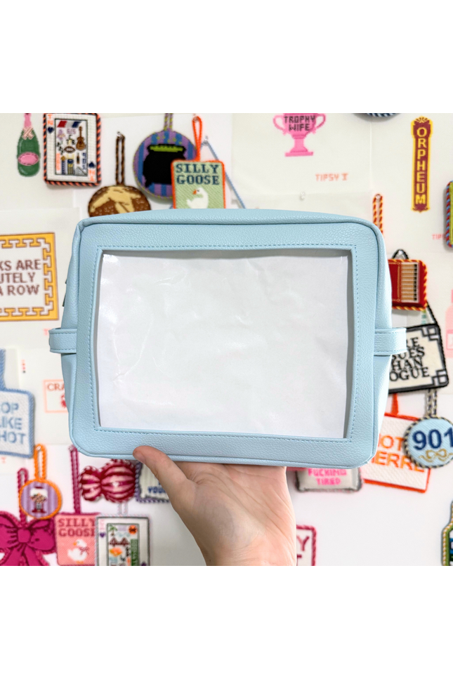 Light blue self finishing needlepoint pouch held by a hand against a colorful background with various small objects and text.