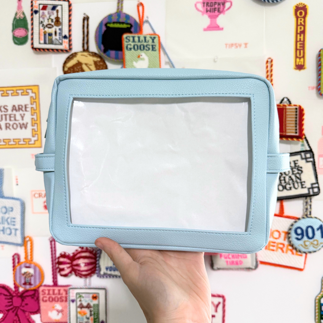 Light blue self finishing needlepoint pouch held by a hand against a colorful background with various small objects and text.