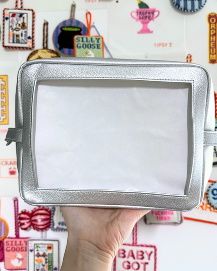 Silver self-finishing needlepoint pouch held in front of a colorful background with various text and graphics.