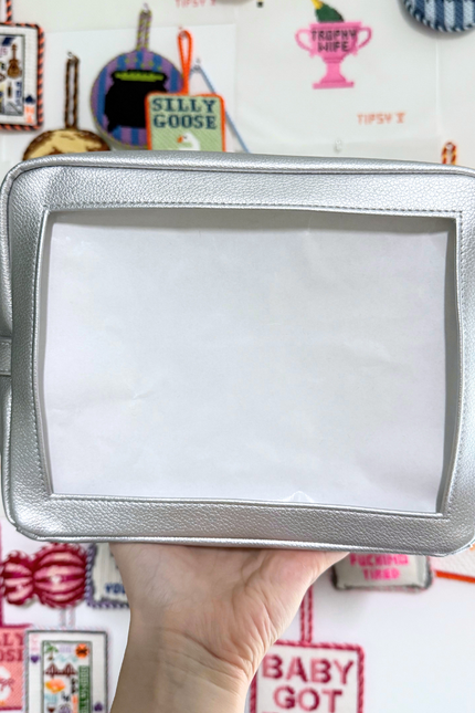 Silver self-finishing needlepoint pouch held in front of a colorful background with various text and graphics.