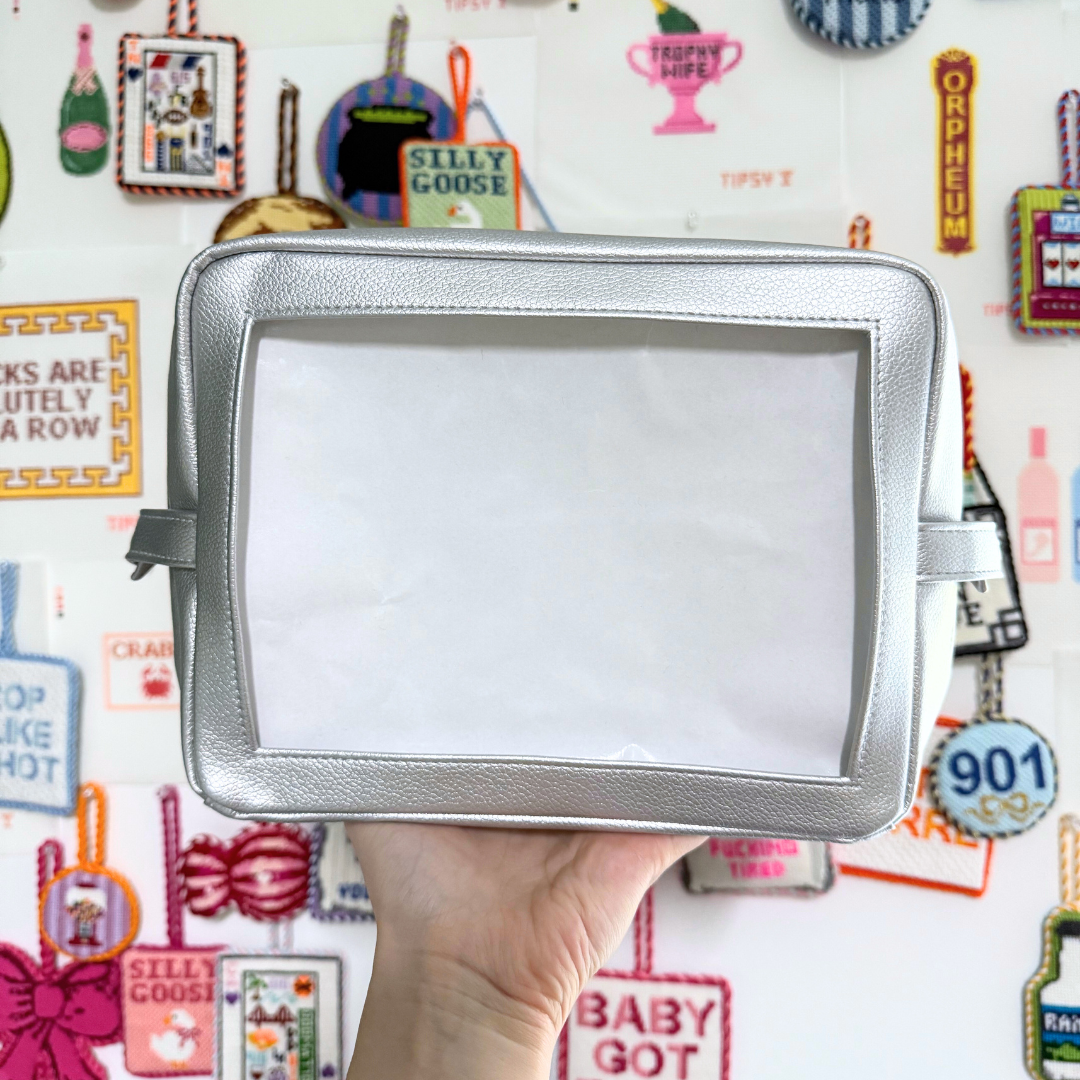 Silver self-finishing needlepoint pouch held in front of a colorful background with various text and graphics.