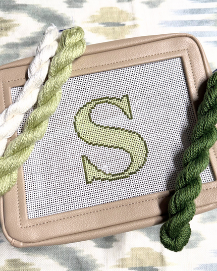 needlepoint kit with a completed letter 'S' design on canvas, surrounded by green yarn on a patterned background.