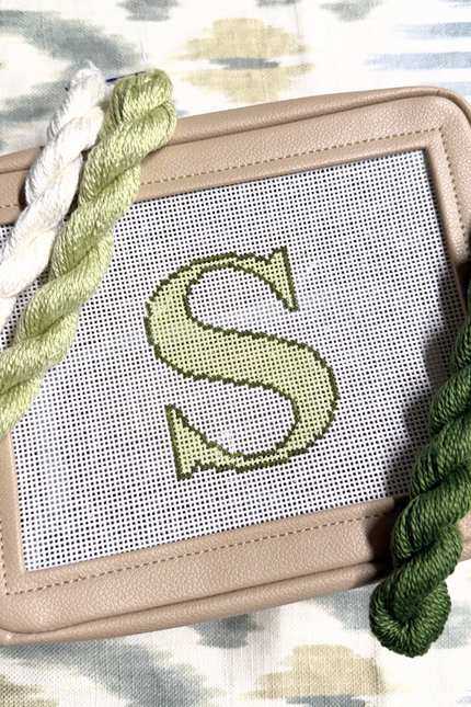 needlepoint kit with a completed letter 'S' design on canvas, surrounded by green yarn on a patterned background.