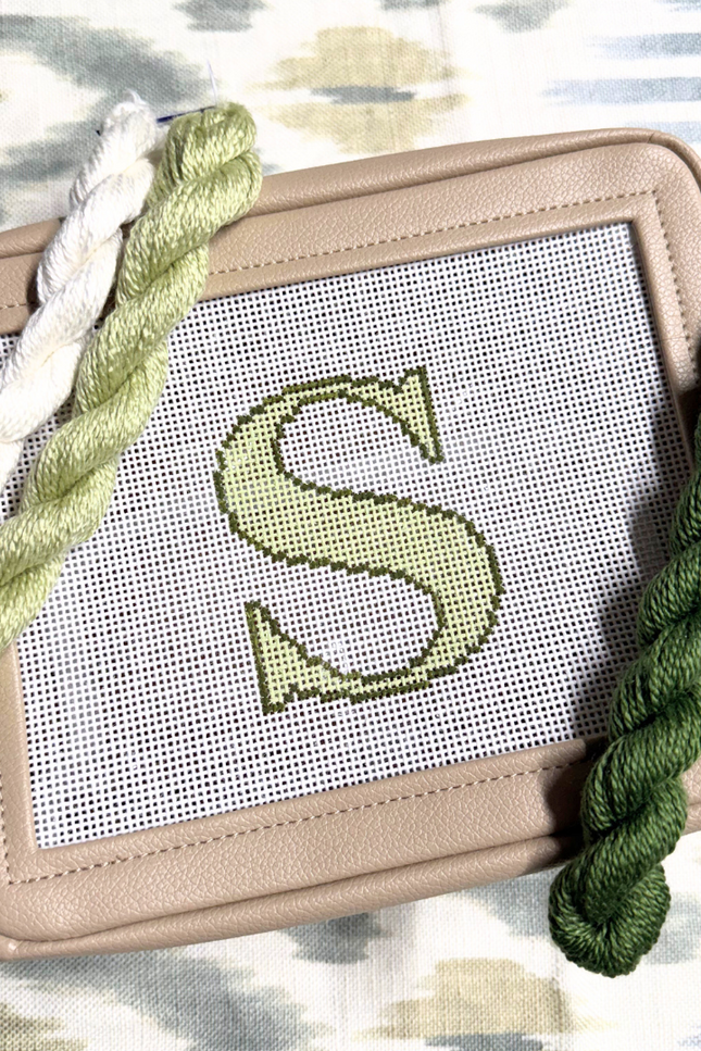 needlepoint kit with a completed letter 'S' design on canvas, surrounded by green yarn on a patterned background.