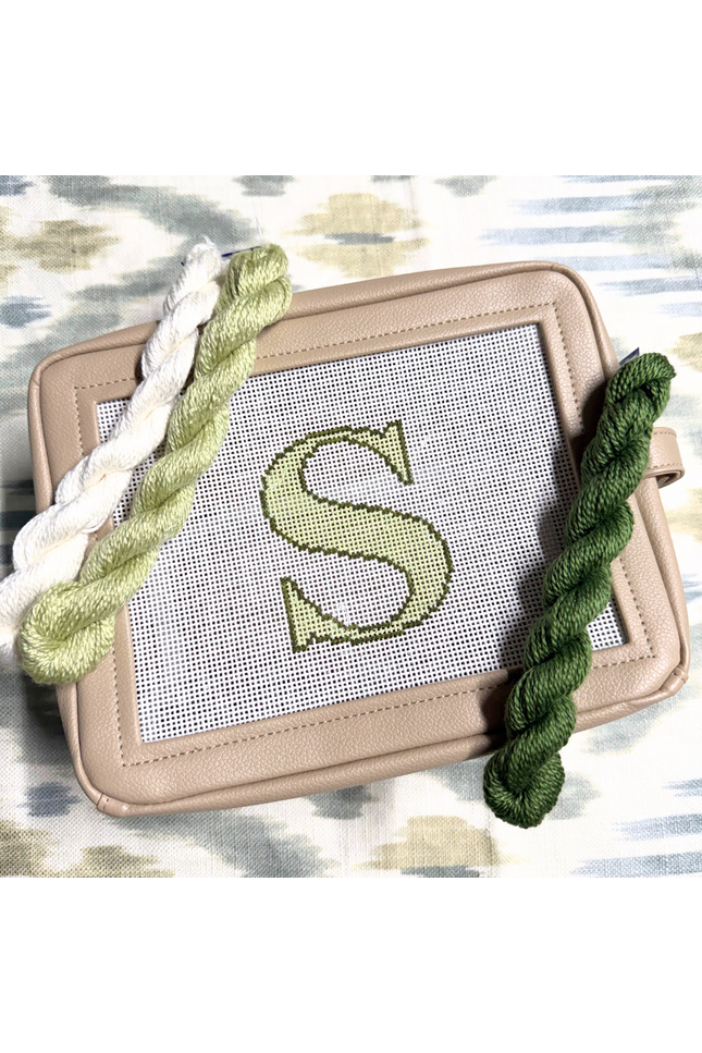 needlepoint kit with a completed letter 'S' design on canvas, surrounded by green yarn on a patterned background.